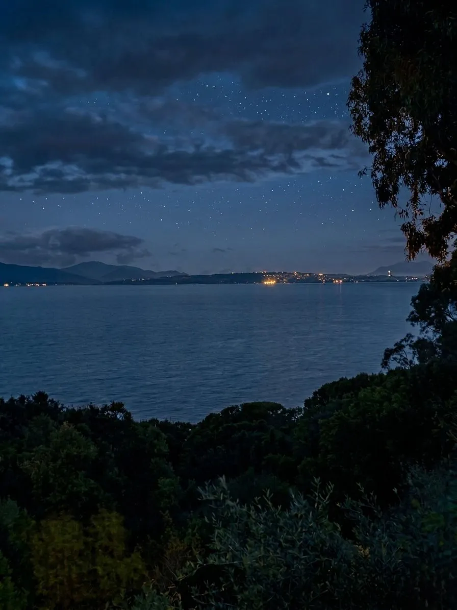 Vista notturna dal balcone della suite a Cagliari: cielo stellato sul Golfo, le luci di Cagliari all'orizzonte — da Marina Piccola al Castello — riflesse sul mare calmo, silhouette della vegetazione mediterranea in primo piano.